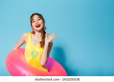 Image of a beautiful excited happy young pin up pretty summer girl in swimwear posing isolated over blue background in inflatable ring showing okay.