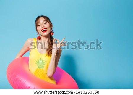 Image of a beautiful excited happy young pin up pretty summer girl in swimwear posing isolated over blue background in inflatable ring showing okay.