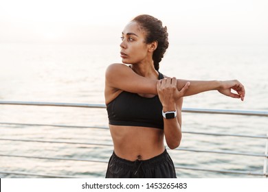 Image of beautiful fitness woman in sportive clothes stretching her arms while doing workout by seaside in morning - Powered by Shutterstock