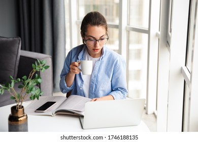 Image of beautiful focused woman drinking tea and working with laptop while sitting at table in living room - Shutterstock ID 1746188135