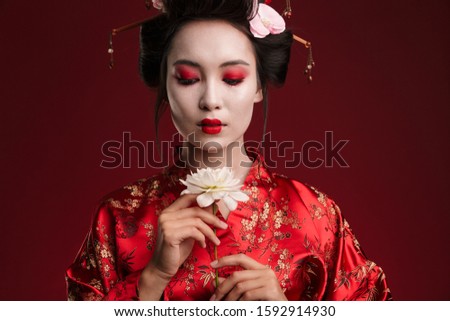 Image of beautiful geisha woman in traditional japanese kimono holding flower isolated over red background