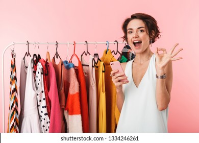 Image Of A Beautiful Happy Woman Stylist Posing Isolated Over Pink Wall Background Near A Lot Of Clothes Using Mobile Phone.