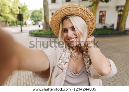 Image of beautiful lady wearing suit and straw hat smiling while taking selfie photo outdoor in downtown