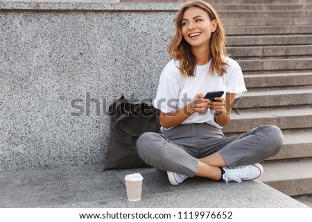 Image of beautiful stylish woman sitting on street stairs with legs crossed on summer day and holding mobile phone