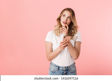 Image of a beautiful thinking young blonde woman posing isolated over pink wall background using mobile phone.