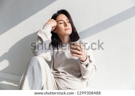 Image of beautiful woman 30s holding cup with tea while sitting over white wall indoor