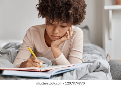Image of beautiful woman with Afro hairstyle rewrites information in notebook from book, writes down homework, enjoys leisure time in bedroom, wears casual clothes. Female makes future plans