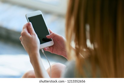 Image Of A Beautiful Woman Sitting On The Bed, Looking At The Phone And Listening To Music On Headphones After Waking Up With A Feeling Of Happiness.