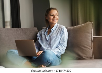 Image of beautiful young joyful woman using laptop and smiling while sitting on sofa at living room - Shutterstock ID 1718474230