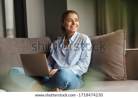 Image of beautiful young joyful woman using laptop and smiling while sitting on sofa at living room