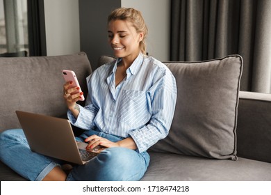 Image of beautiful young smiling woman using laptop and talking on cellphone while sitting on sofa at home