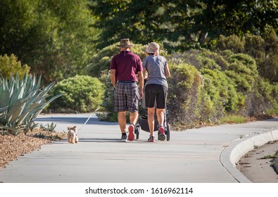 Image From Behind Older Couple With Man Wearing Plaid Pants Pushing Stroller And Walking Thier Dog Down A Sidewalk