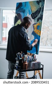 Image of a black man painting on an easel inside of his apartment. He is patiently and slowly applying colour. Window view on the building next door.