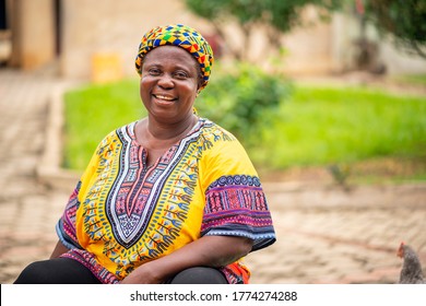 Image Of Black Mother With Beautiful African Print Dress And Head Wrapper-African Woman Smiling In Her Garden-black Woman Sitting On A Chair With Joy And Happiness.