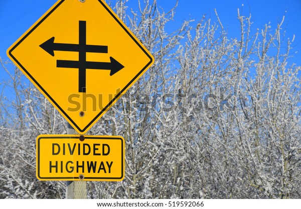 An image of a black and yellow divided highway\
sign in winter.