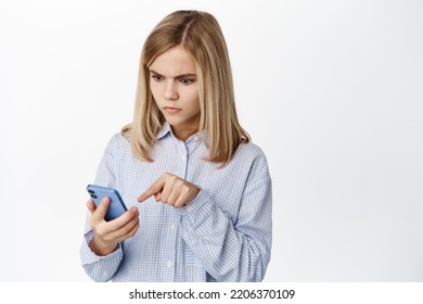 Image Of Blond Little Kid, Girl Staring At Smartphone With Shocked Confused Face, Standing Over White Background.