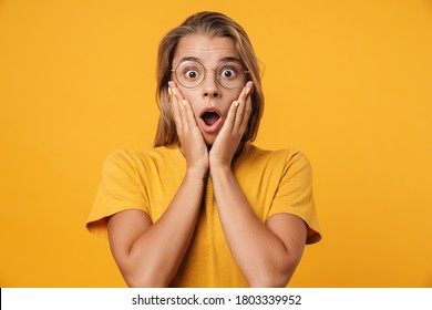 Image of blonde shocked woman in eyeglasses expressing surprise on camera isolated over yellow background