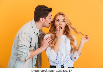 Image of brunette man whispering secret or interesting gossip to surprised woman in her ear isolated over yellow background
