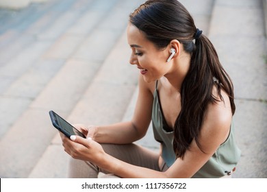 Image of brunette young woman with dark hair in ponytail sitting on street stairs and holding mobile phone while listening to music via bluetooth earpod