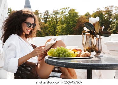 Image of brunette young woman wearing sunglasses drinking coffee and reading book on terrace outdoors