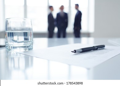 Image of business document, pen and glass of water at workplace with group of colleagues on background 