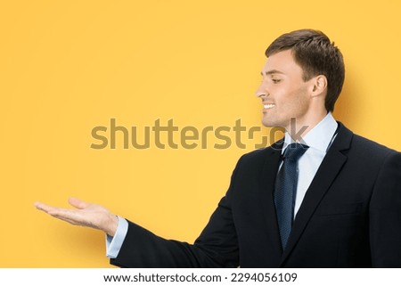 Image of business man in black confident suit, necktie, hold, show, giving advertising product, isolated orange yellow background. Copy space, slogan text area. Advertisement ad concept. Profile view