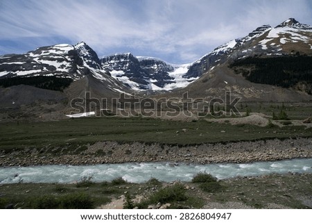 The image captures a broad panoramic vista of the Snow Dome glacier icefall descending into a vast subalpine valley in Jasper National Park, Alberta. The center background features the massive, flat-topped peak with its sweeping white ice cap, flanked by steep, snow-streaked mountain walls on both sides. A wide, turquoise glacial meltwater river flows rapidly across the rocky, gravel-strewn foreground basin under a bright sky with wispy clouds.