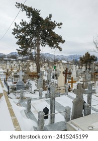 The image captures a cemetery with marble crosses, accompanied by a fir tree, with mountains rising in the background. The landscape conveys harmony between past and nature