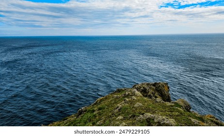 The image captures the cliff's edge with rocks and green grass in the foreground, leading to a calm sea in beautiful shades of blue under a cloudy sky, creating a sense of the edge of the world.