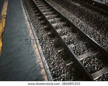 The image captures a close-up, high-angle view of a railway track running parallel to a station platform. The sturdy steel rails rest firmly on concrete sleepers, securely anchored over a dense bed of grey ballast stones that fill the trackbed. On the left side, the edge of the concrete platform features a worn yellow safety line, showing textures of age and continuous public use. The dramatic contrast between the highlighted metallic rails and the deep shadows among the gravel creates a moody, industrial atmosphere, evoking themes of transit, journeys, and daily commuting.