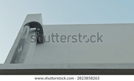 The image captures a close-up, low-angle shot of a white concrete wall against a clear, light blue sky. On the left side, a vertical concrete pillar supports a gray PVC pipe assembly, which includes two 90-degree elbow joints creating an inverted U-shape. The pipe appears weathered with visible scuffs, and it extends downward through a horizontal ledge that runs across the lower portion of the frame. The overall composition is minimalist, focusing on the geometric lines of the structure and the functional plumbing element.