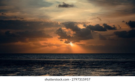 The image captures a dramatic coastal landscape during a vibrant sunset or sunrise, composed with a wide, eye-level perspective that emphasizes the vastness of the scene. The background is dominated by a sweeping sky filled with heavy, dark grey and charcoal clouds that are dynamically pierced by the bright, fiery light of the sun near the centre. This intense light creates prominent, angled sunbeams—crepuscular rays—that fan downwards toward the horizon, bathing the surrounding clouds in rich shades of amber, vivid orange, and muted gold. Below the straight, distinct horizon line, the middle ground features a dark, expansive body of water with a subtly rippled surface that reflects the ambient warmth of the sky. In the immediate foreground, a dark, uneven expanse of wet tidal flats or mudflats grounds the image; this area is mostly cast in deep shadow but is intricately textured with shallow pools of water that catch fragmented, horizontal reflections of the sunset above. The distinct contrasting textures between the soft, voluminous clouds, the gently churning water, and the slick, textured mud contribute to a profoundly moody, tranquil, and atmospheric composition.
Location : Kutch ( Gujarat , India )