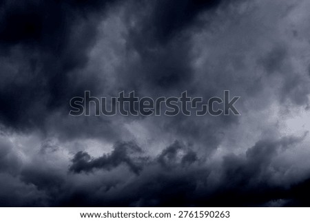 The image captures a dramatic expanse of dark, turbulent storm clouds filling the entire frame. There is no visible horizon, land, or human-made structure—only the sky, heavy and brooding, as if on the verge of unleashing rain or thunder. The clouds vary in density and tone, ranging from deep charcoal and navy hues to lighter grays that glow faintly where light filters through.At the top of the image, the clouds are especially dense and dark, forming a thick canopy that feels weighty and oppressive. A lighter band of cloud stretches horizontally across the middle of the frame, acting as a visual break between the darker masses above and below.