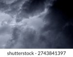 The image captures a dramatic expanse of sky dominated by towering storm clouds. Thick, heavy masses of dark gray and charcoal clouds loom from both sides of the frame, curving inward as if closing around a brighter opening at the center. The clouds appear dense and textured, with soft, billowing edges that suggest constant movement and turbulence. Their forms are layered and sculpted, with deeper shadows pooling in the folds and lighter grays tracing the contours where faint light filters through.
At the center of the composition, a pale, diffused glow breaks through the gloom. This lighter patch of sky, almost silvery in tone, creates a striking contrast against the surrounding darkness. It feels like a temporary clearing, a narrow corridor of light between two advancing walls of cloud. The brightness does not come from direct sunlight but rather from sunlight obscured by thick vapor, giving the scene a moody, atmospheric quality.
The left side of the image shows clouds with slightly more variation in tone, where subtle gradations of gray transition from smoky light to near-black. The right side appears heavier and darker, with a more imposing, shadowed presence. The cloud formations rise upward and inward, giving the impression of height and scale, as though the viewer is looking up 
