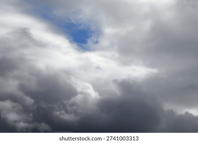 The image captures a dramatic expanse of sky dominated by towering storm clouds. Thick, heavy masses of dark gray and charcoal clouds loom from both sides of the frame, curving inward as if closing around a brighter opening at the center. The clouds appear dense and textured, with soft, billowing edges that suggest constant movement and turbulence. Their forms are layered and sculpted, with deeper shadows pooling in the folds and lighter grays tracing the contours where faint light filters through.
At the center of the composition, a pale, diffused glow breaks through the gloom. This lighter patch of sky, almost silvery in tone, creates a striking contrast against the surrounding darkness. It feels like a temporary clearing, a narrow corridor of light between two advancing walls of cloud. The brightness does not come from direct sunlight but rather from sunlight obscured by thick vapor, giving the scene a moody, atmospheric quality.
The left side of the image shows clouds with slightly more variation in tone, where subtle gradations of gray transition from smoky light to near-black. The right side appears heavier and darker, with a more imposing, shadowed presence. The cloud formations rise upward and inward, giving the impression of height and scale, as though the viewer is looking up from the ground at a vast, shifting ceiling of weather.
There are no visible landmarks, buildings, or horizon lines—only sky. This absence of grounding elements enhances the feeling of immersion, as if the viewer is enveloped by the storm. The image conveys tension and anticipation, suggesting that rain or thunder may soon follow. At the same time, the central glow hints at resilience and contrast—light pushing gently against encroaching darkness. The overall mood is powerful, contemplative, and slightly ominous, capturing the raw, ever-changing drama of the atmosphere.