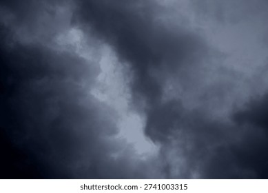 The image captures a dramatic expanse of sky dominated by towering storm clouds. Thick, heavy masses of dark gray and charcoal clouds loom from both sides of the frame, curving inward as if closing around a brighter opening at the center. The clouds appear dense and textured, with soft, billowing edges that suggest constant movement and turbulence. Their forms are layered and sculpted, with deeper shadows pooling in the folds and lighter grays tracing the contours where faint light filters through.
At the center of the composition, a pale, diffused glow breaks through the gloom. This lighter patch of sky, almost silvery in tone, creates a striking contrast against the surrounding darkness. It feels like a temporary clearing, a narrow corridor of light between two advancing walls of cloud. The brightness does not come from direct sunlight but rather from sunlight obscured by thick vapor, giving the scene a moody, atmospheric quality.
The left side of the image shows clouds with slightly more variation in tone, where subtle gradations of gray transition from smoky light to near-black. The right side appears heavier and darker, with a more imposing, shadowed presence. The cloud formations rise upward and inward, giving the impression of height and scale, as though the viewer is looking up from the ground at a vast, shifting ceiling of weather.
There are no visible landmarks, buildings, or horizon lines—only sky. This absence of grounding elements enhances the feeling of immersion, as if the viewer is enveloped by the storm. The image conveys tension and anticipation, suggesting that rain or thunder may soon follow. At the same time, the central glow hints at resilience and contrast—light pushing gently against encroaching darkness. The overall mood is powerful, contemplative, and slightly ominous, capturing the raw, ever-changing drama of the atmosphere.
