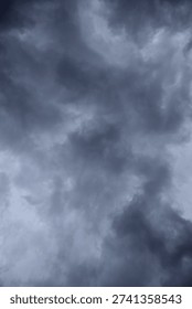 The image captures a dramatic expanse of sky dominated by towering storm clouds. Thick, heavy masses of dark gray and charcoal clouds loom from both sides of the frame, curving inward as if closing around a brighter opening at the center. The clouds appear dense and textured, with soft, billowing edges that suggest constant movement and turbulence. Their forms are layered and sculpted, with deeper shadows pooling in the folds and lighter grays tracing the contours where faint light filters through.
At the center of the composition, a pale, diffused glow breaks through the gloom. This lighter patch of sky, almost silvery in tone, creates a striking contrast against the surrounding darkness. It feels like a temporary clearing, a narrow corridor of light between two advancing walls of cloud. The brightness does not come from direct sunlight but rather from sunlight obscured by thick vapor, giving the scene a moody, atmospheric quality.
The left side of the image shows clouds with slightly more variation in tone, where subtle gradations of gray transition from smoky light to near-black. The right side appears heavier and darker, with a more imposing, shadowed presence. The cloud formations rise upward and inward, giving the impression of height and scale, as though the viewer is looking up from the ground at a vast, shifting ceiling of weather.
There are no visible landmarks, buildings, or horizon lines—only sky. This absence of grounding elements enhances the feeling of immersion, as if the viewer is enveloped by the storm. The image conveys tension and anticipation, suggesting that rain or thunder may soon follow. At the same time, the central glow hints at resilience and contrast—light pushing gently against encroaching darkness. The overall mood is powerful, contemplative, and slightly ominous, capturing the raw, ever-changing drama of the atmosphere.