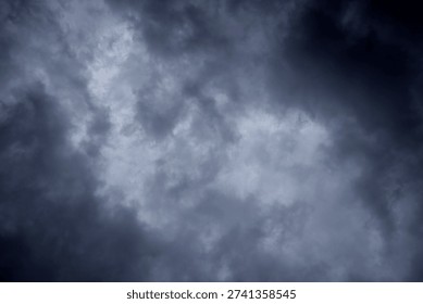 The image captures a dramatic expanse of sky dominated by towering storm clouds. Thick, heavy masses of dark gray and charcoal clouds loom from both sides of the frame, curving inward as if closing around a brighter opening at the center. The clouds appear dense and textured, with soft, billowing edges that suggest constant movement and turbulence. Their forms are layered and sculpted, with deeper shadows pooling in the folds and lighter grays tracing the contours where faint light filters through.
At the center of the composition, a pale, diffused glow breaks through the gloom. This lighter patch of sky, almost silvery in tone, creates a striking contrast against the surrounding darkness. It feels like a temporary clearing, a narrow corridor of light between two advancing walls of cloud. The brightness does not come from direct sunlight but rather from sunlight obscured by thick vapor, giving the scene a moody, atmospheric quality.
The left side of the image shows clouds with slightly more variation in tone, where subtle gradations of gray transition from smoky light to near-black. The right side appears heavier and darker, with a more imposing, shadowed presence. The cloud formations rise upward and inward, giving the impression of height and scale, as though the viewer is looking up from the ground at a vast, shifting ceiling of weather.
There are no visible landmarks, buildings, or horizon lines—only sky. This absence of grounding elements enhances the feeling of immersion, as if the viewer is enveloped by the storm. The image conveys tension and anticipation, suggesting that rain or thunder may soon follow. At the same time, the central glow hints at resilience and contrast—light pushing gently against encroaching darkness. The overall mood is powerful, contemplative, and slightly ominous, capturing the raw, ever-changing drama of the atmosphere.