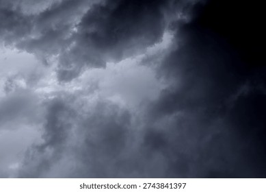 The image captures a dramatic expanse of sky dominated by towering storm clouds. Thick, heavy masses of dark gray and charcoal clouds loom from both sides of the frame, curving inward as if closing around a brighter opening at the center. The clouds appear dense and textured, with soft, billowing edges that suggest constant movement and turbulence. Their forms are layered and sculpted, with deeper shadows pooling in the folds and lighter grays tracing the contours where faint light filters through.
At the center of the composition, a pale, diffused glow breaks through the gloom. This lighter patch of sky, almost silvery in tone, creates a striking contrast against the surrounding darkness. It feels like a temporary clearing, a narrow corridor of light between two advancing walls of cloud. The brightness does not come from direct sunlight but rather from sunlight obscured by thick vapor, giving the scene a moody, atmospheric quality.
The left side of the image shows clouds with slightly more variation in tone, where subtle gradations of gray transition from smoky light to near-black. The right side appears heavier and darker, with a more imposing, shadowed presence. The cloud formations rise upward and inward, giving the impression of height and scale, as though the viewer is looking up 