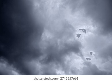 The image captures a dramatic expanse of sky dominated by towering storm clouds. Thick, heavy masses of dark gray and charcoal clouds loom from both sides of the frame, curving inward as if closing around a brighter opening at the center. The clouds appear dense and textured, with soft, billowing edges that suggest constant movement and turbulence. Their forms are layered and sculpted, with deeper shadows pooling in the folds and lighter grays tracing the contours where faint light filters through.
At the center of the composition, a pale, diffused glow breaks through the gloom. This lighter patch of sky, almost silvery in tone, creates a striking contrast against the surrounding darkness. It feels like a temporary clearing, a narrow corridor of light between two advancing walls of cloud. The brightness does not come from direct sunlight but rather from sunlight obscured by thick vapor, giving the scene a moody, atmospheric quality.
The left side of the image shows clouds with slightly more variation in tone, where subtle gradations of gray transition from smoky light to near-black. The right side appears heavier and darker, with a more imposing, shadowed presence. The cloud formations rise upward and inward, giving the impression of height and scale, as though the viewer is looking up from the ground at a vast, shifting ceiling of weather.
There are no visible landmarks, buildings, or horizon lines—only sky. This absence of grounding elements enhances the feeling of immersion, as if the viewer is enveloped by the storm. The image conveys tension and anticipation, suggesting that rain or thunder may soon follow. At the same time, the central glow hints at resilience and contrast—light pushing gently against encroaching darkness. The overall mood is powerful, contemplative, and slightly ominous, capturing the raw, ever-changing drama of the atmosphere.