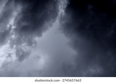 The image captures a dramatic expanse of sky dominated by towering storm clouds. Thick, heavy masses of dark gray and charcoal clouds loom from both sides of the frame, curving inward as if closing around a brighter opening at the center. The clouds appear dense and textured, with soft, billowing edges that suggest constant movement and turbulence. Their forms are layered and sculpted, with deeper shadows pooling in the folds and lighter grays tracing the contours where faint light filters through.
At the center of the composition, a pale, diffused glow breaks through the gloom. This lighter patch of sky, almost silvery in tone, creates a striking contrast against the surrounding darkness. It feels like a temporary clearing, a narrow corridor of light between two advancing walls of cloud. The brightness does not come from direct sunlight but rather from sunlight obscured by thick vapor, giving the scene a moody, atmospheric quality.
The left side of the image shows clouds with slightly more variation in tone, where subtle gradations of gray transition from smoky light to near-black. The right side appears heavier and darker, with a more imposing, shadowed presence. The cloud formations rise upward and inward, giving the impression of height and scale, as though the viewer is looking up from the ground at a vast, shifting ceiling of weather.
There are no visible landmarks, buildings, or horizon lines—only sky. This absence of grounding elements enhances the feeling of immersion, as if the viewer is enveloped by the storm. The image conveys tension and anticipation, suggesting that rain or thunder may soon follow. At the same time, the central glow hints at resilience and contrast—light pushing gently against encroaching darkness. The overall mood is powerful, contemplative, and slightly ominous, capturing the raw, ever-changing drama of the atmosphere.