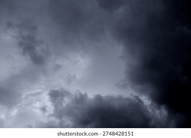 The image captures a dramatic expanse of sky dominated by towering storm clouds. Thick, heavy masses of dark gray and charcoal clouds loom from both sides of the frame, curving inward as if closing around a brighter opening at the center. The clouds appear dense and textured, with soft, billowing edges that suggest constant movement and turbulence. Their forms are layered and sculpted, with deeper shadows pooling in the folds and lighter grays tracing the contours where faint light filters through.
At the center of the composition, a pale, diffused glow breaks through the gloom. This lighter patch of sky, almost silvery in tone, creates a striking contrast against the surrounding darkness. It feels like a temporary clearing, a narrow corridor of light between two advancing walls of cloud. The brightness does not come from direct sunlight but rather from sunlight obscured by thick vapor, giving the scene a moody, atmospheric quality.
The left side of the image shows clouds with slightly more variation in tone, where subtle gradations of gray transition from smoky light to near-black. The right side appears heavier and darker, with a more imposing, shadowed presence. The cloud formations rise upward and inward, giving the impression of height and scale, as though the viewer is looking up from the ground at a vast, shifting ceiling of weather.
There are no visible landmarks, buildings, or horizon lines—only sky. This absence of grounding elements enhances the feeling of immersion, as if the viewer is enveloped by the storm. The image conveys tension and anticipation, suggesting that rain or thunder may soon follow. At the same time, the central glow hints at resilience and contrast—light pushing gently against encroaching darkness. The overall mood is powerful, contemplative, and slightly ominous, capturing the raw, ever-changing drama of the atmosphere.