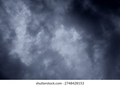 The image captures a dramatic expanse of sky dominated by towering storm clouds. Thick, heavy masses of dark gray and charcoal clouds loom from both sides of the frame, curving inward as if closing around a brighter opening at the center. The clouds appear dense and textured, with soft, billowing edges that suggest constant movement and turbulence. Their forms are layered and sculpted, with deeper shadows pooling in the folds and lighter grays tracing the contours where faint light filters through.
At the center of the composition, a pale, diffused glow breaks through the gloom. This lighter patch of sky, almost silvery in tone, creates a striking contrast against the surrounding darkness. It feels like a temporary clearing, a narrow corridor of light between two advancing walls of cloud. The brightness does not come from direct sunlight but rather from sunlight obscured by thick vapor, giving the scene a moody, atmospheric quality.
The left side of the image shows clouds with slightly more variation in tone, where subtle gradations of gray transition from smoky light to near-black. The right side appears heavier and darker, with a more imposing, shadowed presence. The cloud formations rise upward and inward, giving the impression of height and scale, as though the viewer is looking up from the ground at a vast, shifting ceiling of weather.
There are no visible landmarks, buildings, or horizon lines—only sky. This absence of grounding elements enhances the feeling of immersion, as if the viewer is enveloped by the storm. The image conveys tension and anticipation, suggesting that rain or thunder may soon follow. At the same time, the central glow hints at resilience and contrast—light pushing gently against encroaching darkness. The overall mood is powerful, contemplative, and slightly ominous, capturing the raw, ever-changing drama of the atmosphere.