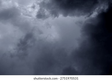 The image captures a dramatic expanse of sky dominated by towering storm clouds. Thick, heavy masses of dark gray and charcoal clouds loom from both sides of the frame, curving inward as if closing around a brighter opening at the center. The clouds appear dense and textured, with soft, billowing edges that suggest constant movement and turbulence. Their forms are layered and sculpted, with deeper shadows pooling in the folds and lighter grays tracing the contours where faint light filters through.
At the center of the composition, a pale, diffused glow breaks through the gloom. This lighter patch of sky, almost silvery in tone, creates a striking contrast against the surrounding darkness. It feels like a temporary clearing, a narrow corridor of light between two advancing walls of cloud. The brightness does not come from direct sunlight but rather from sunlight obscured by thick vapor, giving the scene a moody, atmospheric quality.
The left side of the image shows clouds with slightly more variation in tone, where subtle gradations of gray transition from smoky light to near-black. The right side appears heavier and darker, with a more imposing, shadowed presence. The cloud formations rise upward and inward, giving the impression of height and scale, as though the viewer is looking up from the ground at a vast, shifting ceiling of weather.
There are no visible landmarks, buildings, or horizon lines—only sky. This absence of grounding elements enhances the feeling of immersion, as if the viewer is enveloped by the storm. The image conveys tension and anticipation, suggesting that rain or thunder may soon follow. At the same time, the central glow hints at resilience and contrast—light pushing gently against encroaching darkness. The overall mood is powerful, contemplative, and slightly ominous, capturing the raw, ever-changing drama of the atmosphere.