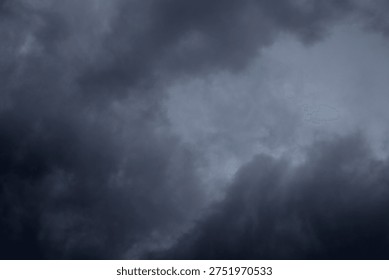 The image captures a dramatic expanse of sky dominated by towering storm clouds. Thick, heavy masses of dark gray and charcoal clouds loom from both sides of the frame, curving inward as if closing around a brighter opening at the center. The clouds appear dense and textured, with soft, billowing edges that suggest constant movement and turbulence. Their forms are layered and sculpted, with deeper shadows pooling in the folds and lighter grays tracing the contours where faint light filters through.
At the center of the composition, a pale, diffused glow breaks through the gloom. This lighter patch of sky, almost silvery in tone, creates a striking contrast against the surrounding darkness. It feels like a temporary clearing, a narrow corridor of light between two advancing walls of cloud. The brightness does not come from direct sunlight but rather from sunlight obscured by thick vapor, giving the scene a moody, atmospheric quality.
The left side of the image shows clouds with slightly more variation in tone, where subtle gradations of gray transition from smoky light to near-black. The right side appears heavier and darker, with a more imposing, shadowed presence. The cloud formations rise upward and inward, giving the impression of height and scale, as though the viewer is looking up from the ground at a vast, shifting ceiling of weather.
There are no visible landmarks, buildings, or horizon lines—only sky. This absence of grounding elements enhances the feeling of immersion, as if the viewer is enveloped by the storm. The image conveys tension and anticipation, suggesting that rain or thunder may soon follow. At the same time, the central glow hints at resilience and contrast—light pushing gently against encroaching darkness. The overall mood is powerful, contemplative, and slightly ominous, capturing the raw, ever-changing drama of the atmosphere.