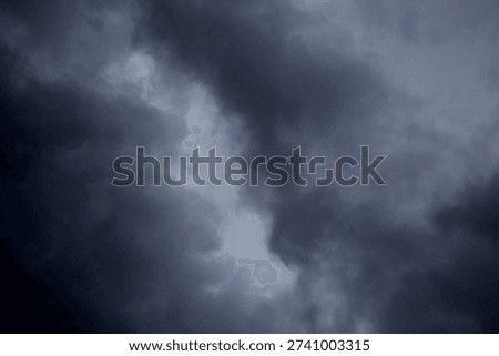 The image captures a dramatic expanse of sky dominated by towering storm clouds. Thick, heavy masses of dark gray and charcoal clouds loom from both sides of the frame, curving inward as if closing around a brighter opening at the center. The clouds appear dense and textured, with soft, billowing edges that suggest constant movement and turbulence. Their forms are layered and sculpted, with deeper shadows pooling in the folds and lighter grays tracing the contours where faint light filters through.
At the center of the composition, a pale, diffused glow breaks through the gloom. This lighter patch of sky, almost silvery in tone, creates a striking contrast against the surrounding darkness. It feels like a temporary clearing, a narrow corridor of light between two advancing walls of cloud. The brightness does not come from direct sunlight but rather from sunlight obscured by thick vapor, giving the scene a moody, atmospheric quality.
The left side of the image shows clouds with slightly more variation in tone, where subtle gradations of gray transition from smoky light to near-black. The right side appears heavier and darker, with a more imposing, shadowed presence. The cloud formations rise upward and inward, giving the impression of height and scale, as though the viewer is looking up from the ground at a vast, shifting ceiling of weather.
There are no visible landmarks, buildings, or horizon lines—only sky. This absence of grounding elements enhances the feeling of immersion, as if the viewer is enveloped by the storm. The image conveys tension and anticipation, suggesting that rain or thunder may soon follow. At the same time, the central glow hints at resilience and contrast—light pushing gently against encroaching darkness. The overall mood is powerful, contemplative, and slightly ominous, capturing the raw, ever-changing drama of the atmosphere.