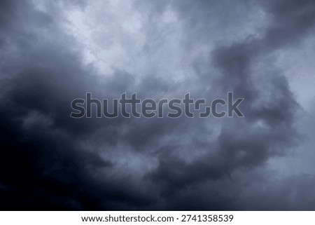 The image captures a dramatic expanse of sky dominated by towering storm clouds. Thick, heavy masses of dark gray and charcoal clouds loom from both sides of the frame, curving inward as if closing around a brighter opening at the center. The clouds appear dense and textured, with soft, billowing edges that suggest constant movement and turbulence. Their forms are layered and sculpted, with deeper shadows pooling in the folds and lighter grays tracing the contours where faint light filters through.
At the center of the composition, a pale, diffused glow breaks through the gloom. This lighter patch of sky, almost silvery in tone, creates a striking contrast against the surrounding darkness. It feels like a temporary clearing, a narrow corridor of light between two advancing walls of cloud. The brightness does not come from direct sunlight but rather from sunlight obscured by thick vapor, giving the scene a moody, atmospheric quality.
The left side of the image shows clouds with slightly more variation in tone, where subtle gradations of gray transition from smoky light to near-black. The right side appears heavier and darker, with a more imposing, shadowed presence. The cloud formations rise upward and inward, giving the impression of height and scale, as though the viewer is looking up from the ground at a vast, shifting ceiling of weather.
There are no visible landmarks, buildings, or horizon lines—only sky. This absence of grounding elements enhances the feeling of immersion, as if the viewer is enveloped by the storm. The image conveys tension and anticipation, suggesting that rain or thunder may soon follow. At the same time, the central glow hints at resilience and contrast—light pushing gently against encroaching darkness. The overall mood is powerful, contemplative, and slightly ominous, capturing the raw, ever-changing drama of the atmosphere.