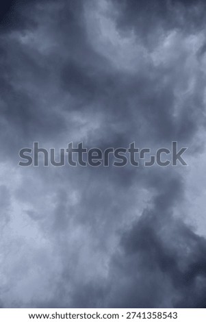 The image captures a dramatic expanse of sky dominated by towering storm clouds. Thick, heavy masses of dark gray and charcoal clouds loom from both sides of the frame, curving inward as if closing around a brighter opening at the center. The clouds appear dense and textured, with soft, billowing edges that suggest constant movement and turbulence. Their forms are layered and sculpted, with deeper shadows pooling in the folds and lighter grays tracing the contours where faint light filters through.
At the center of the composition, a pale, diffused glow breaks through the gloom. This lighter patch of sky, almost silvery in tone, creates a striking contrast against the surrounding darkness. It feels like a temporary clearing, a narrow corridor of light between two advancing walls of cloud. The brightness does not come from direct sunlight but rather from sunlight obscured by thick vapor, giving the scene a moody, atmospheric quality.
The left side of the image shows clouds with slightly more variation in tone, where subtle gradations of gray transition from smoky light to near-black. The right side appears heavier and darker, with a more imposing, shadowed presence. The cloud formations rise upward and inward, giving the impression of height and scale, as though the viewer is looking up from the ground at a vast, shifting ceiling of weather.
There are no visible landmarks, buildings, or horizon lines—only sky. This absence of grounding elements enhances the feeling of immersion, as if the viewer is enveloped by the storm. The image conveys tension and anticipation, suggesting that rain or thunder may soon follow. At the same time, the central glow hints at resilience and contrast—light pushing gently against encroaching darkness. The overall mood is powerful, contemplative, and slightly ominous, capturing the raw, ever-changing drama of the atmosphere.