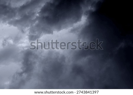 The image captures a dramatic expanse of sky dominated by towering storm clouds. Thick, heavy masses of dark gray and charcoal clouds loom from both sides of the frame, curving inward as if closing around a brighter opening at the center. The clouds appear dense and textured, with soft, billowing edges that suggest constant movement and turbulence. Their forms are layered and sculpted, with deeper shadows pooling in the folds and lighter grays tracing the contours where faint light filters through.
At the center of the composition, a pale, diffused glow breaks through the gloom. This lighter patch of sky, almost silvery in tone, creates a striking contrast against the surrounding darkness. It feels like a temporary clearing, a narrow corridor of light between two advancing walls of cloud. The brightness does not come from direct sunlight but rather from sunlight obscured by thick vapor, giving the scene a moody, atmospheric quality.
The left side of the image shows clouds with slightly more variation in tone, where subtle gradations of gray transition from smoky light to near-black. The right side appears heavier and darker, with a more imposing, shadowed presence. The cloud formations rise upward and inward, giving the impression of height and scale, as though the viewer is looking up 