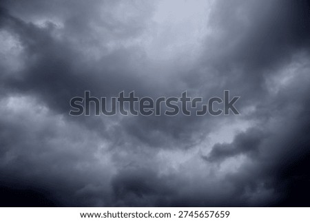 The image captures a dramatic expanse of sky dominated by towering storm clouds. Thick, heavy masses of dark gray and charcoal clouds loom from both sides of the frame, curving inward as if closing around a brighter opening at the center. The clouds appear dense and textured, with soft, billowing edges that suggest constant movement and turbulence. Their forms are layered and sculpted, with deeper shadows pooling in the folds and lighter grays tracing the contours where faint light filters through.
At the center of the composition, a pale, diffused glow breaks through the gloom. This lighter patch of sky, almost silvery in tone, creates a striking contrast against the surrounding darkness. It feels like a temporary clearing, a narrow corridor of light between two advancing walls of cloud. The brightness does not come from direct sunlight but rather from sunlight obscured by thick vapor, giving the scene a moody, atmospheric quality.
The left side of the image shows clouds with slightly more variation in tone, where subtle gradations of gray transition from smoky light to near-black. The right side appears heavier and darker, with a more imposing, shadowed presence. The cloud formations rise upward and inward, giving the impression of height and scale, as though the viewer is looking up from the ground at a vast, shifting ceiling of weather.
There are no visible landmarks, buildings, or horizon lines—only sky. This absence of grounding elements enhances the feeling of immersion, as if the viewer is enveloped by the storm. The image conveys tension and anticipation, suggesting that rain or thunder may soon follow. At the same time, the central glow hints at resilience and contrast—light pushing gently against encroaching darkness. The overall mood is powerful, contemplative, and slightly ominous, capturing the raw, ever-changing drama of the atmosphere.